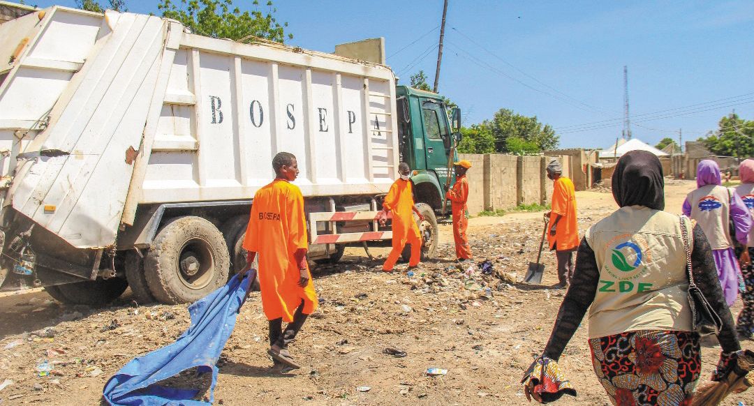 A ZAIDF team member and sanitation workers clear waste beside a BOSEPA refuse truck