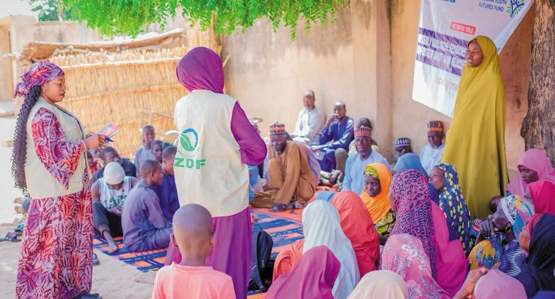 A ZAIDF team member leads a community dialogue session with women and men seated on mats