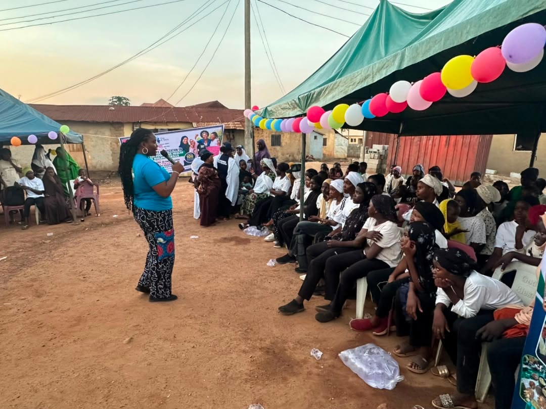 A facilitator speaks to a seated crowd beneath colorful canopies
